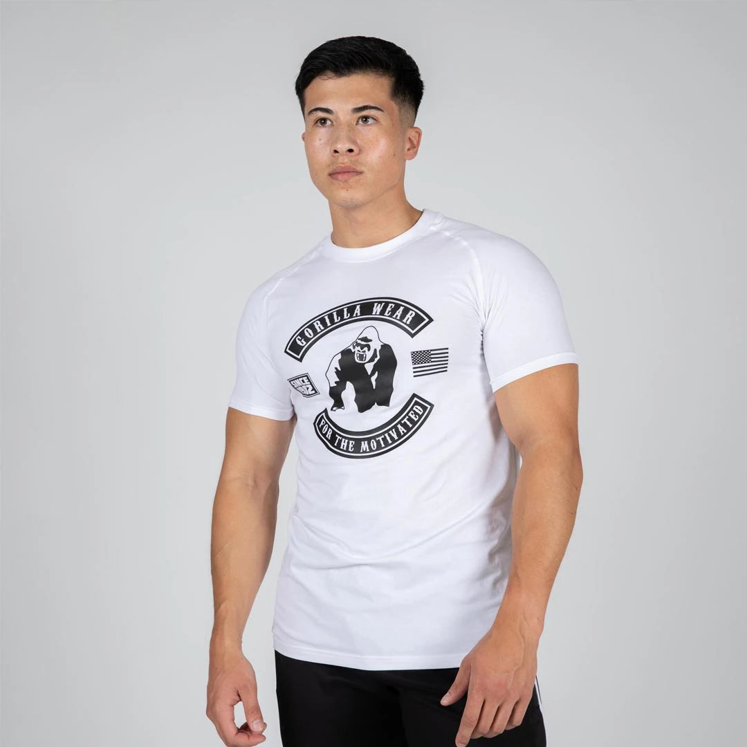 Mann i Gorilla Wear Tulsa T-Shirt White, en hvit kroppsnær trenings-t-skjorte med stort svart logotrykk.