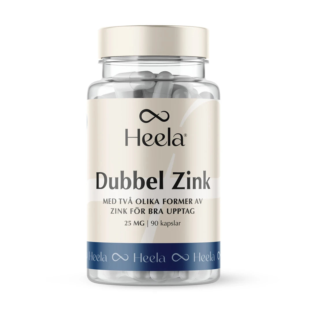 Heela Dubbel Zink 60 caps i en gjennomsiktig boks med hvite kapsler, beigefarget etikett og blå bunnstripe.