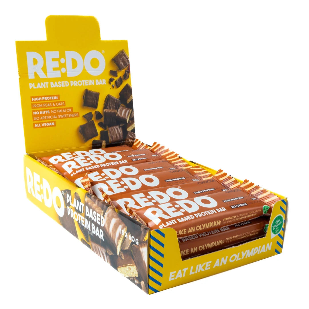 18 x REDO Bar 60 g Chocolate Chock i en åpen gul displayeske fylt med brune proteinbarer.